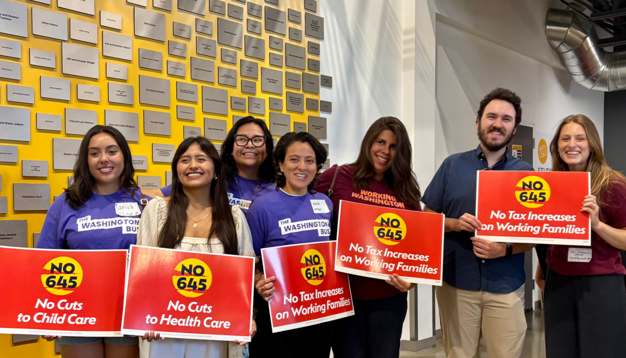 A crowd of Working Washington members and Washington Bus members wearing organizational shirts and holding red signs with white text, reading: No on I-645: No Cuts to Health Care, No Tax Increases on Working Families.